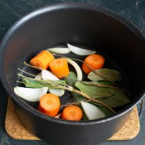 Кладём в кастрюлю луковицу, добавляем морковь, лавровый лист, листья порея, черный перец и корешок петрушки